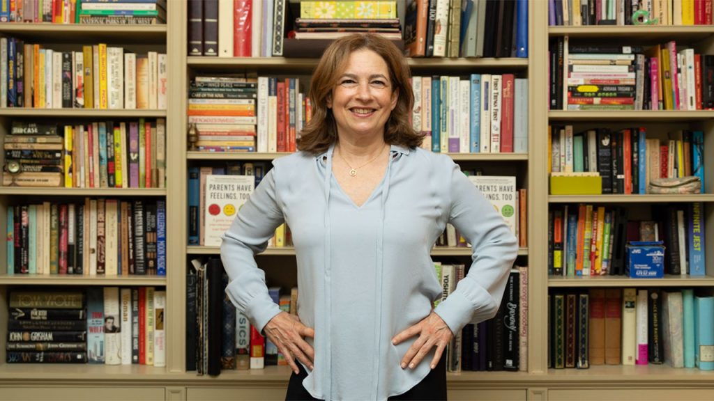 A smiling woman with shoulder-length brown hair, wearing a light blue blouse, standing with her hands on her hips in front of large bookshelves filled with books.