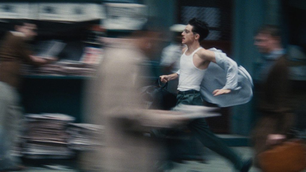 What Makes a Great Screenplay? A still image from Marty Supreme of the protagonist running through the street as other people blur around him.