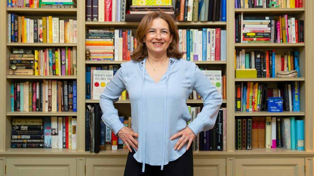 A smiling woman with shoulder-length brown hair, wearing a light blue blouse, standing with her hands on her hips in front of large bookshelves filled with books.