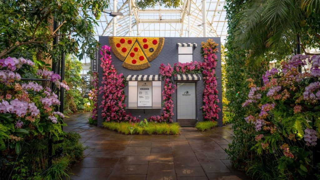 A photo of the inside of the conservatory at the New York Botanical Garden where the Orchid Show is taking place, featuring a building decorated in flowers to look like an NYC pizza shop
