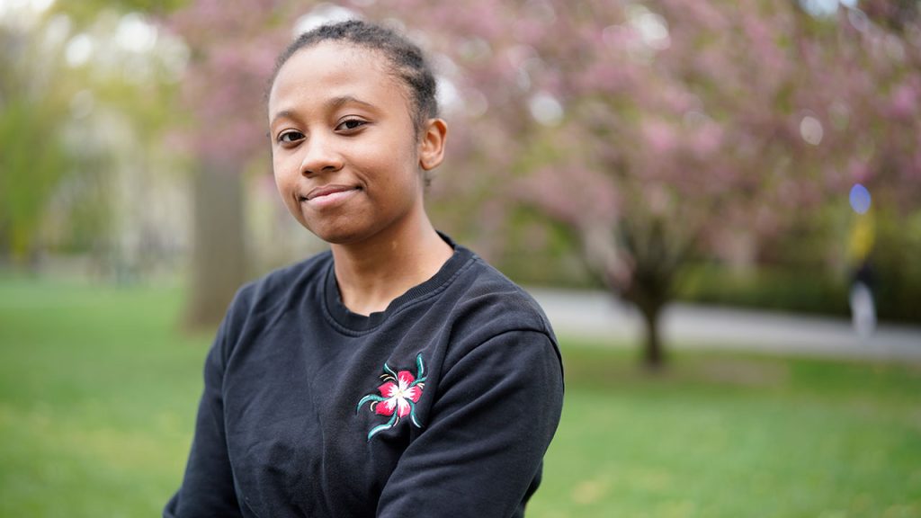 A woman wearing a black long-sleeve shirt with a flower on it smiles for the camera while sitting in front of a flowering tree 