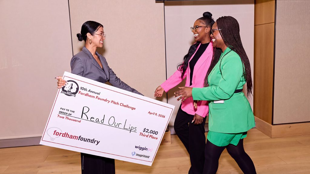 A woman wearing a suit and glasses presents an over-sized check to two women wearing pink and green business suits.