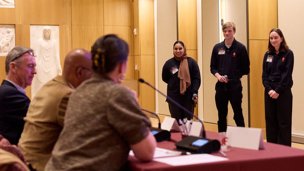 Three students wearing black stand together, listening to a panel of judges speak to them.