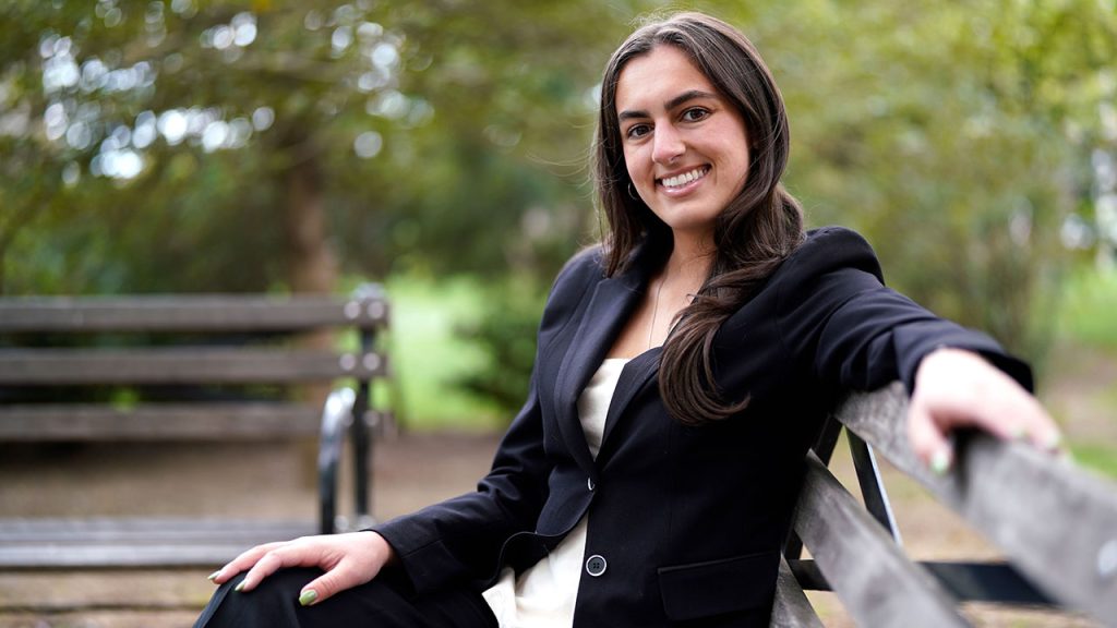 A woman wearing a black suit jacket smiles while seated on a bench, with her arm extended toward the camera