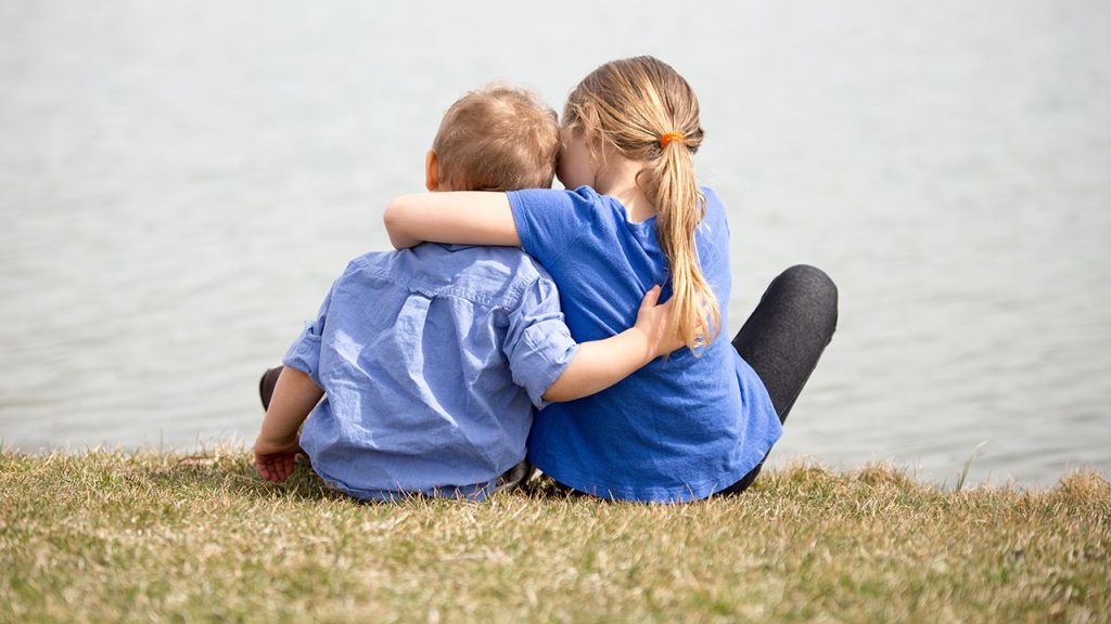 How Do Our Siblings Help Raise Us? Two children, brother and sister, sitting by the water with arms around each other