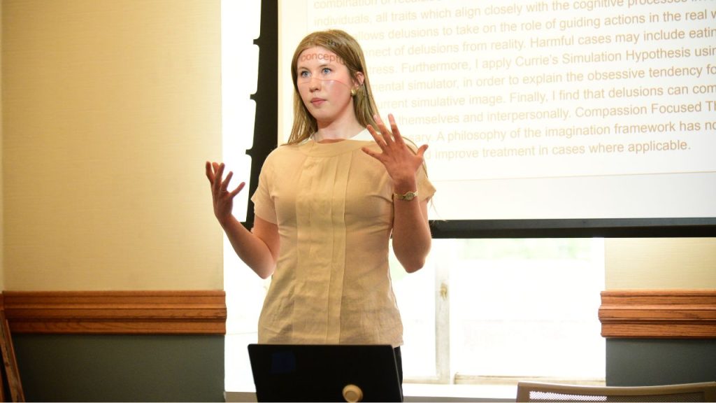 A young woman stands in front of a screen gesturing with both hands 