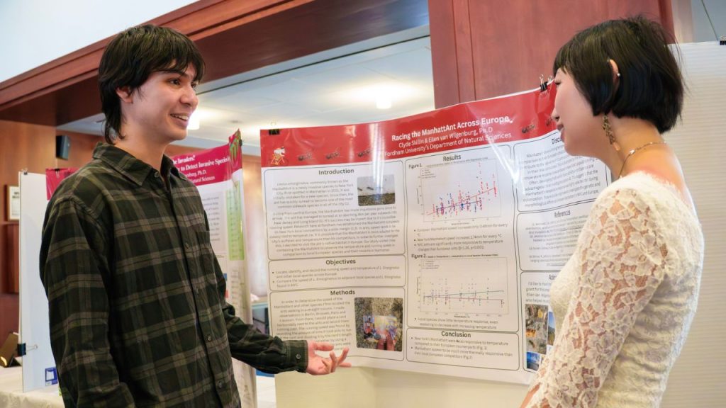 A male student discusses his research with a female student in front of a poster board