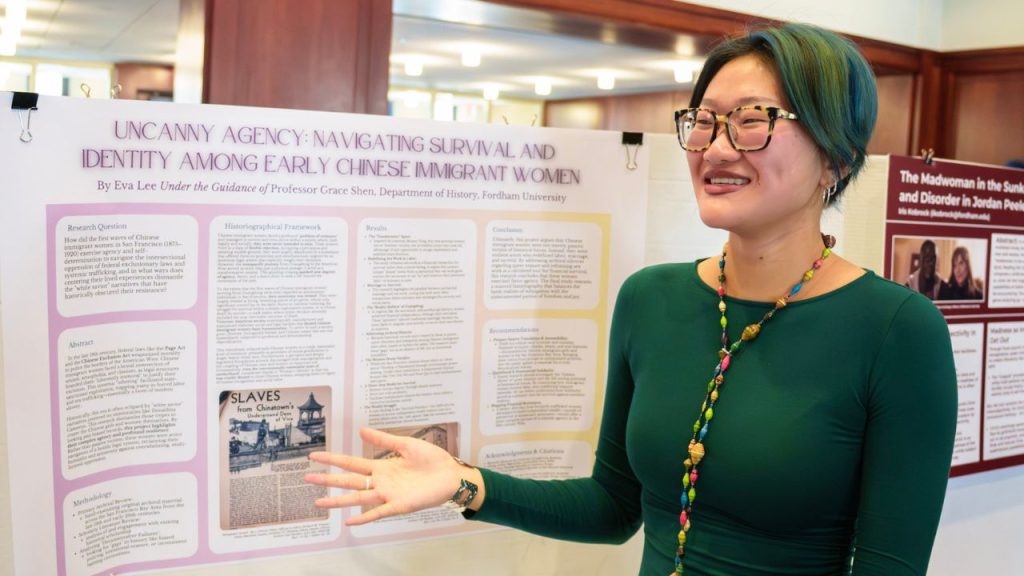A young woman stands in front of a poster board, gesturing and smiling as she discusses her research