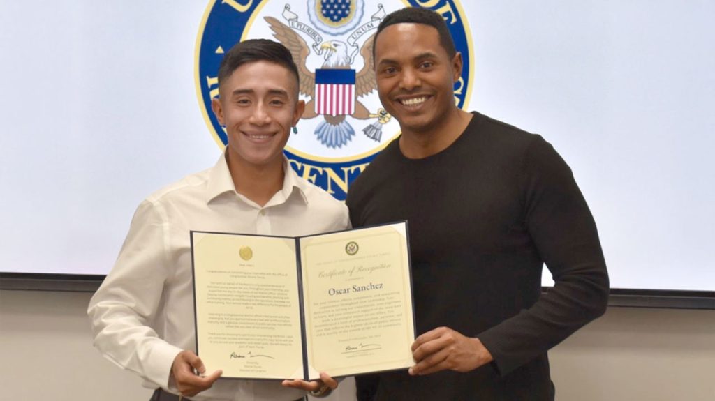 Two men standing together in front of a US seal, one holding a proclamation