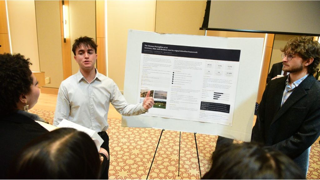 Two young men stand beside a poster board. One speaks to a crowd of onlookers.