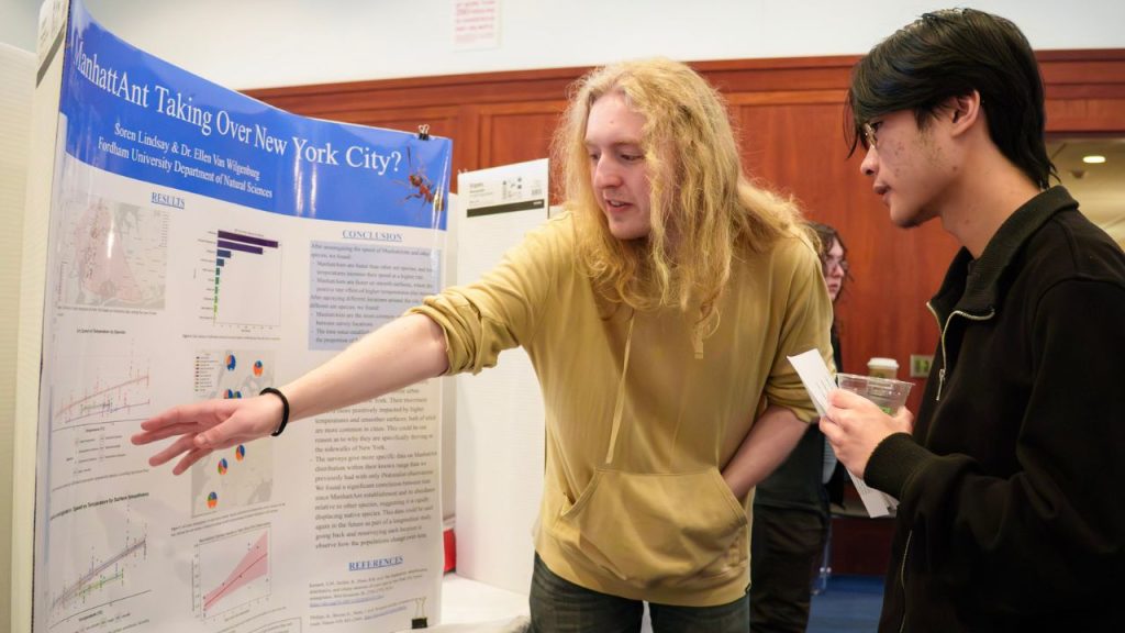 One student points to a poster board while another student listens to him speak.