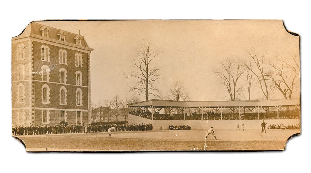 A sepia-toned, panoramic vintage photograph from 1902 showing a baseball game in progress on a dirt and grass field. On the left stands a large, multi-story stone building with arched windows. In the background, a covered wooden grandstand is packed with spectators, and a line of people stands along the field's perimeter.