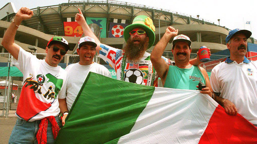Five men cheer outside a stadium during a World Cup match, holding a large Italian flag. The man in the center wears a large green top hat with a shamrock, a shirt decorated with different national flags, sunglasses, and a long beard. The men around him wear casual clothes, sunglasses, and baseball caps, with two raising their fists in celebration. In the background, the concrete stadium features "94" and World Cup soccer ball graphics.