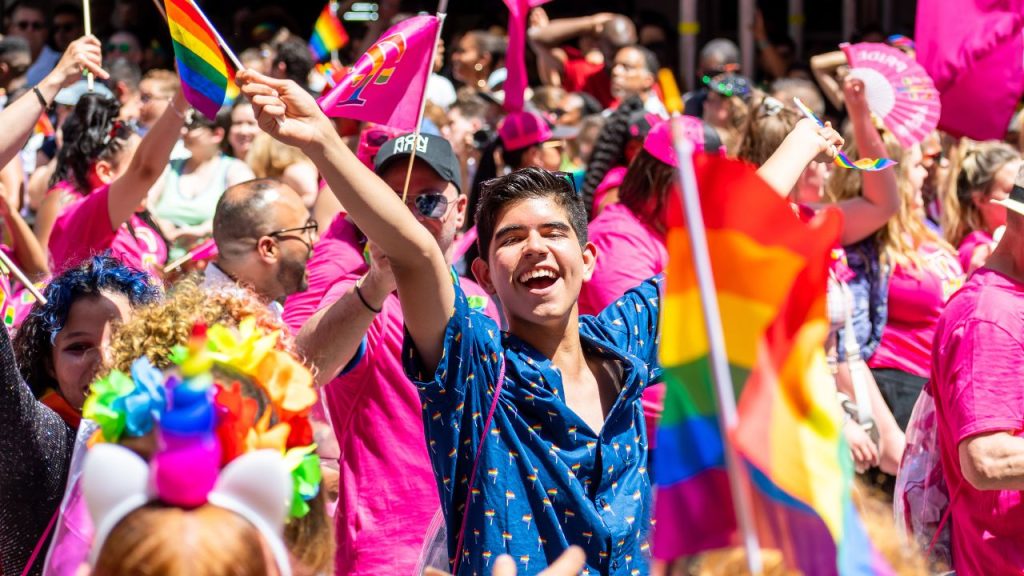A colorful photo of a man waving a rainbow flag in the middle of a crowd at an NYC pride parade