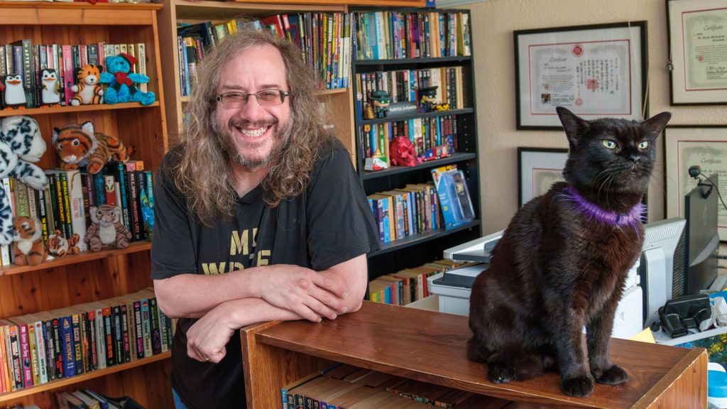 Author Keith R.A. DeCandido smiles warmly while leaning on a wooden shelf in an office filled with packed bookshelves. A sleek black cat wearing a purple fuzzy collar sits upright on the shelf beside him.