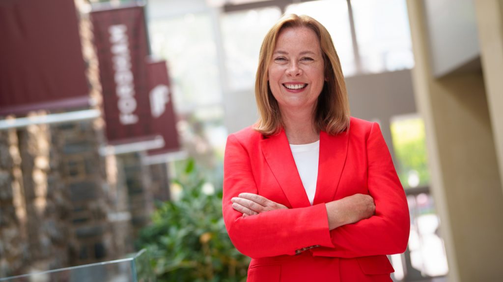 Provost KerryAnn O'Meara wears a bright red blazer and stands and smiles for a portrait in the McShane Center.