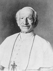 A black-and-white portrait of Pope Leo XIII in white vestments wearing a large, ornate pectoral cross and smiling