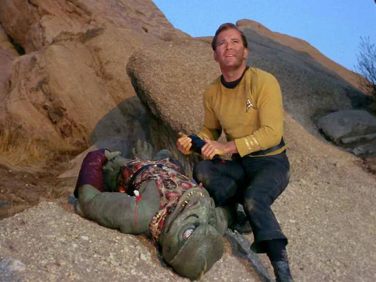 Captain Kirk kneels on a rocky slope holding a improvised weapon, looking upward, while the green, reptilian Gorn alien lies prone beside him among large rock formations in a scene from the original Star Trek episode "Arena."