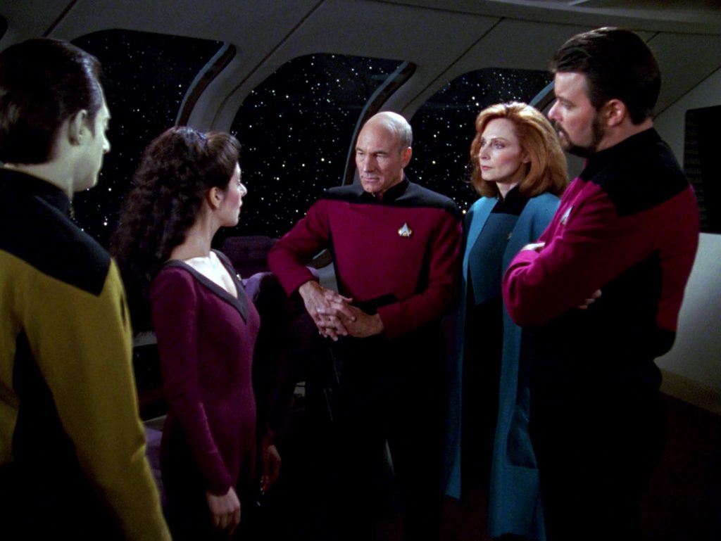 A scene from Star Trek: The Next Generation featuring Data, Deanna Troi, Captain Jean-Luc Picard, Dr. Beverly Crusher, and Commander Riker standing together in a tight circle in the Enterprise observation lounge, engaged in a focused conversation with a starry space view visible through large windows in the background.