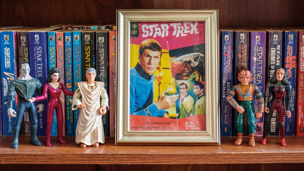 A bookshelf lined with Star Trek paperback novels, a framed classic Gold Key comic book cover featuring Spock, and several Star Trek action figures displayed along the shelf.