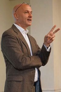 Fordham philosophy professor Stephen Grimm speaking while holding up two fingers, wearing a patterned blazer with eyeglasses rested on his head.