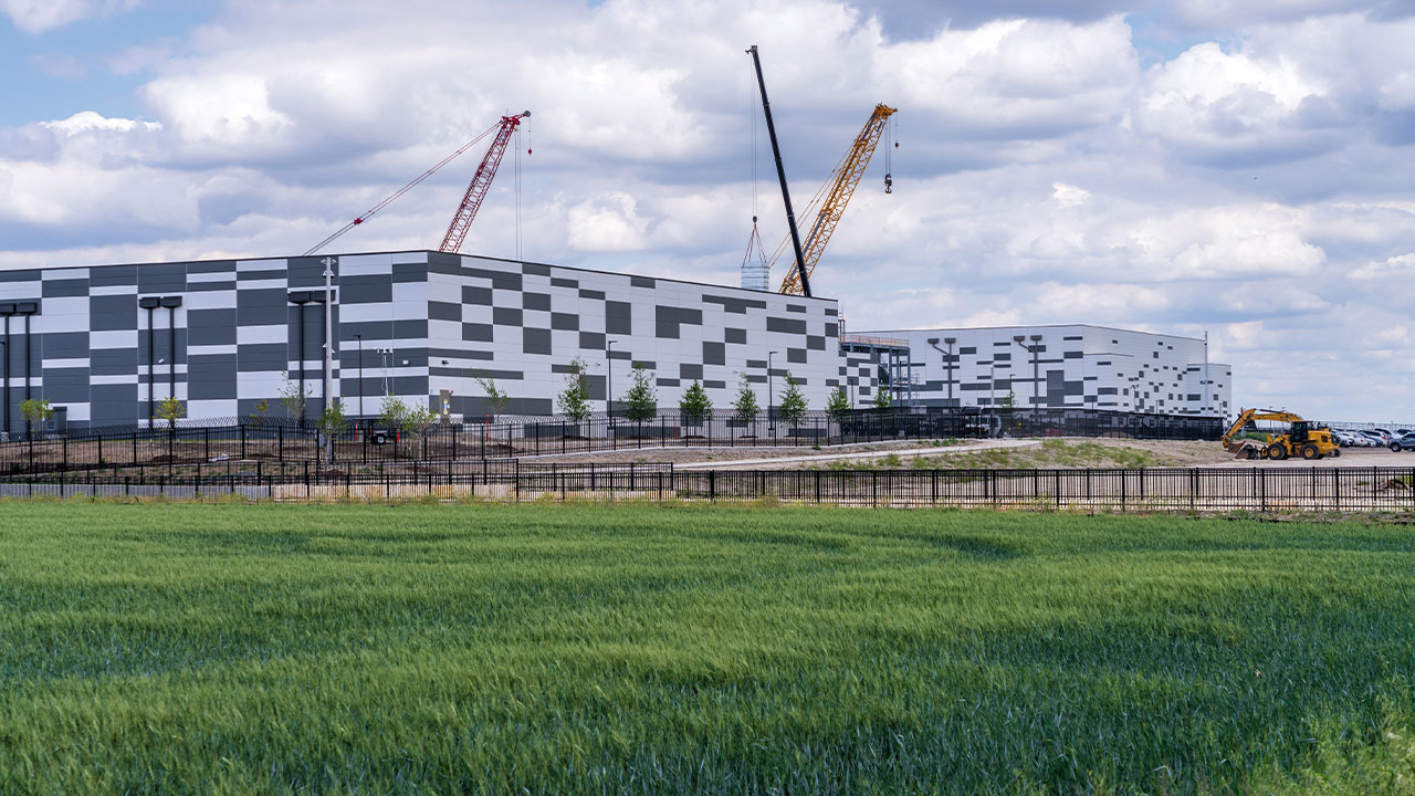 Exterior view of a large, modern data center facility under construction next to a green field, featuring construction cranes and a patterned grey-and-white facade.