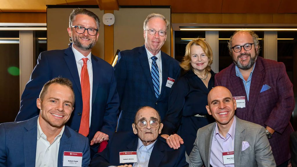 A group of seven people smiling for a photograph at a indoor formal event, with an elderly man seated in the center wearing glasses and a suit, surrounded by six colleagues in business attire