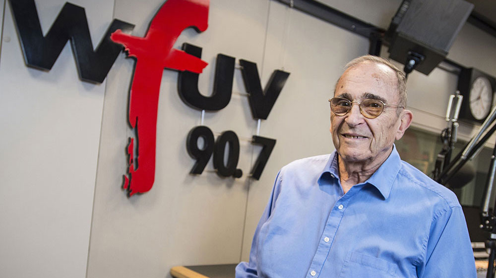 Bob Ahrens, WFUV Sports Director and Beloved Mentor, Dies at 89 A man wearing glasses and a light blue button-up shirt smiling in a radio studio. Behind him, the wall displays the WFUV 90.7 logo.