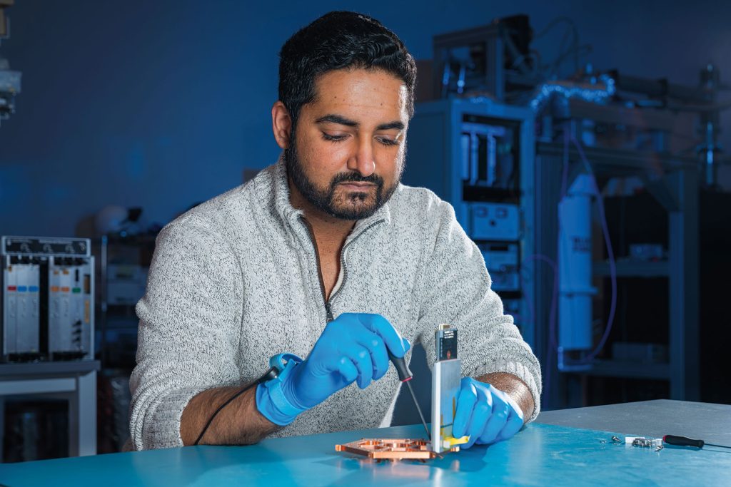 Joshua Sobrin, wearing a light gray sweater and blue nitrile gloves, sits at a lab bench and uses a screwdriver on a copper-colored circular component attached to a circuit board.