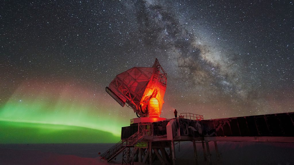 A wide shot of the South Pole Telescope, with its dish and a person standing on its platform illuminated by red light under a starry sky with green aurora