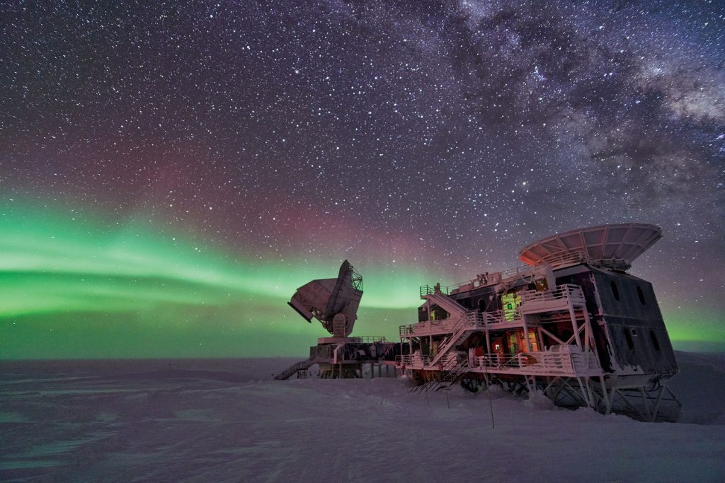 Wide shot of the South Pole Telescope facility and dish against a starry night sky and green aurora