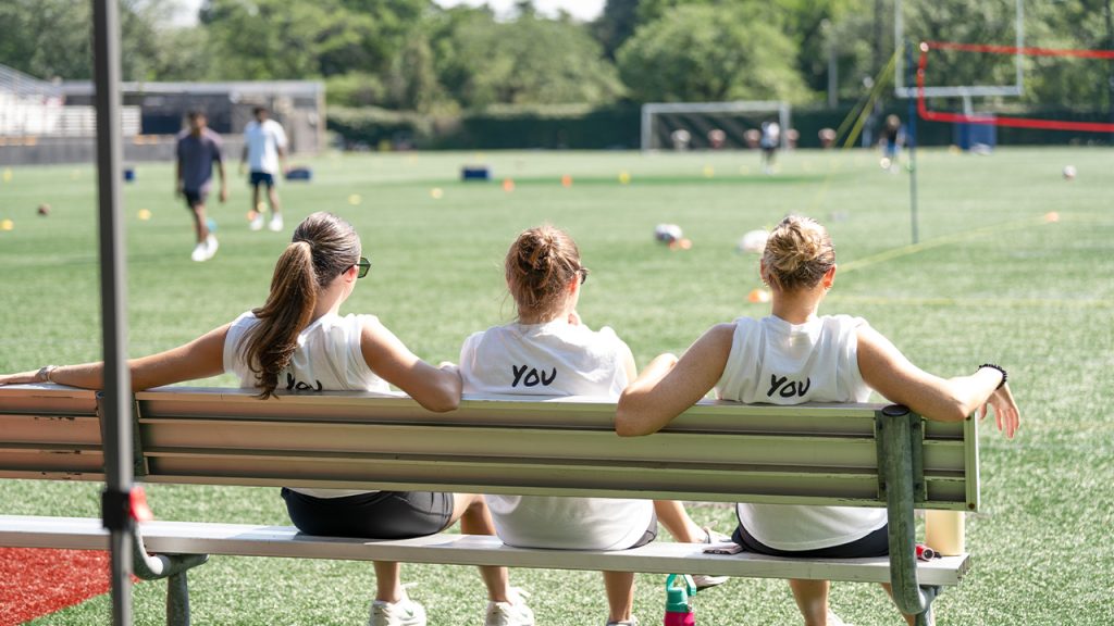 The backs of three people sitting on a bench on the field of Moglia Stadium