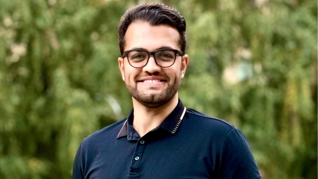 Fordham PhD Student Receives National Psychology Research Award A photo of a man in a navy blue shirt with glasses in front of green foliage.