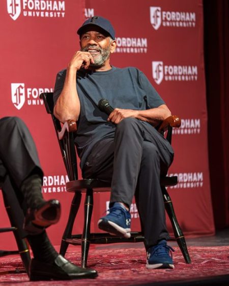 Fordham graduate Denzel Washington sits on a chair onstage in Pope Auditorium, holding a microphone and smiling while wearing a baseball cap and dark casual clothing in front of a maroon Fordham University backdrop.