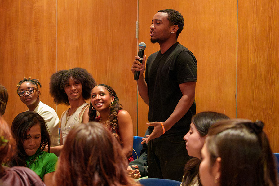 A Fordham Theatre student in a black T-shirt stands and speaks into a microphone during a Q&A session with Denzel Washington and Anthony Carter in Pope Auditorium as fellow students in the audience look on and listen.