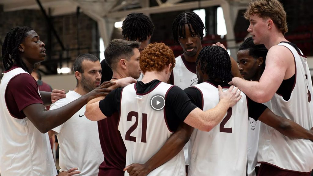 Fordham men's basketball players in a huddle with a video play button