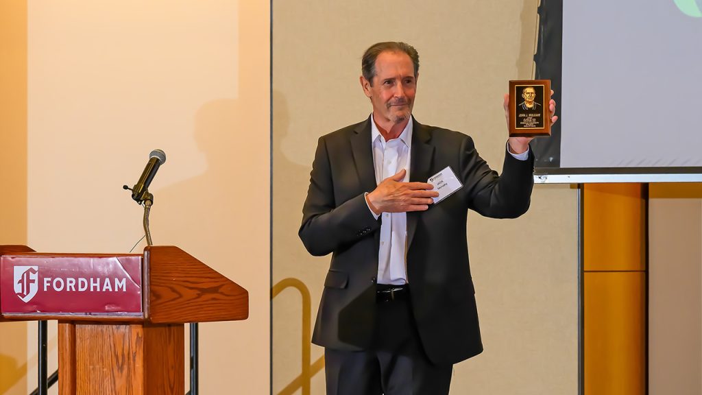 ‘Closing the Loop’ on a Fordham Olympian’s Hall of Fame Honors Jack Mulcahy holding up a plaque honoring his grandfather, John Mulcahy, from the stage at Fordham's Athletic Hall of Fame induction ceremony