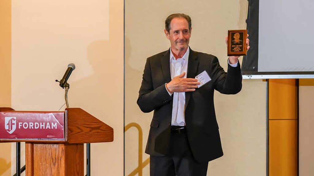 ‘Closing the Loop’ on a Fordham Olympian’s Hall of Fame Honors Jack Mulcahy holding up a plaque honoring his grandfather, John Mulcahy, from the stage at Fordham's Athletic Hall of Fame induction ceremony