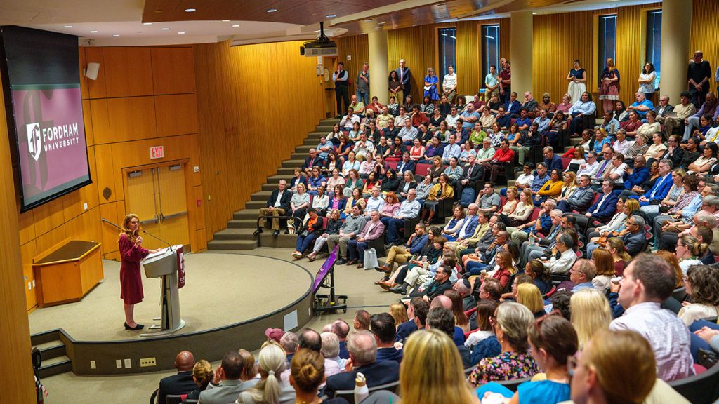 A crowd of seated people gathered in a room watching a woman on the stage