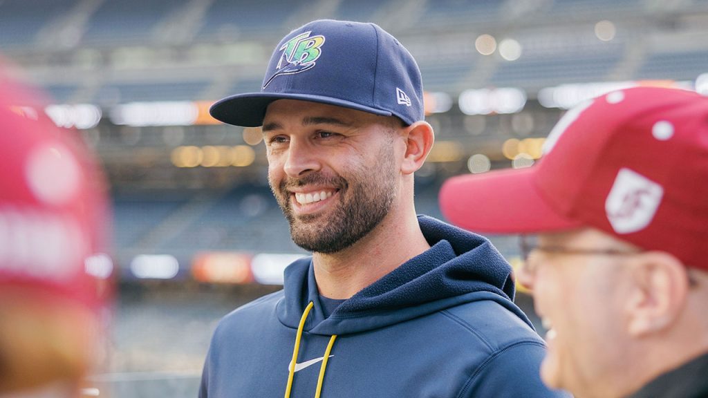 Tampa Bay Rays All-Star Pitcher Nick Martinez, wearing a dark blue Tampa Bay Rays baseball cap and a matching blue hoodie, smiles standing outdoors on the center field deck at Yankee Stadium while talking with fellow Fordham alumni wearing maroon hats.