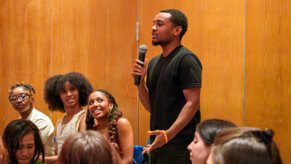 A Fordham Theatre student in a black T-shirt stands and speaks into a microphone during a Q&A session with Denzel Washington and Anthony Carter in Pope Auditorium as fellow students in the audience look on and listen.