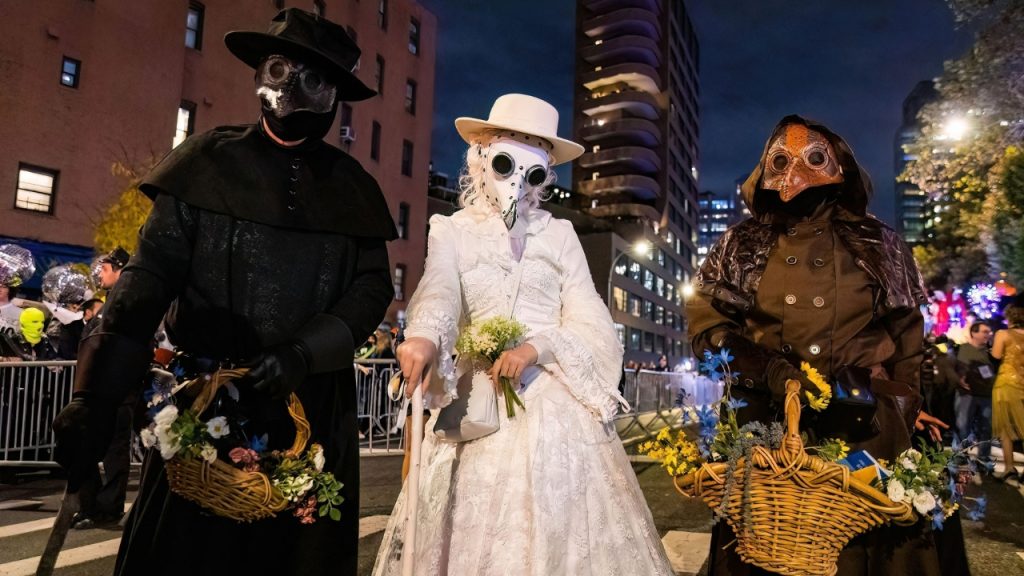 A group of three people in plague doctor masks and costumes, carrying flowers, at the West Village Halloween Parade.