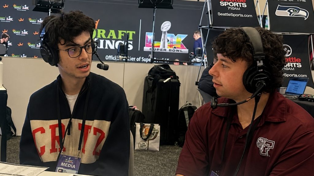 Two young men wearing headsets with microphones chat with each other with Super Bowl branding in the background, representing WFUV's New York State Broadcasting Awards