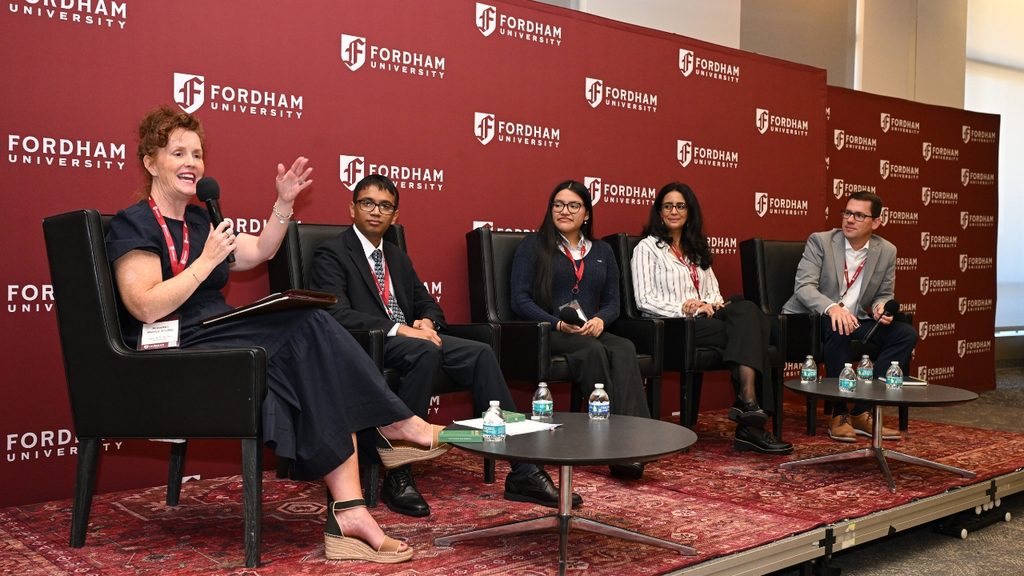 A group of five people sitting in chairs on a stage in front of a Fordham banner at the Ignatian AI Conference