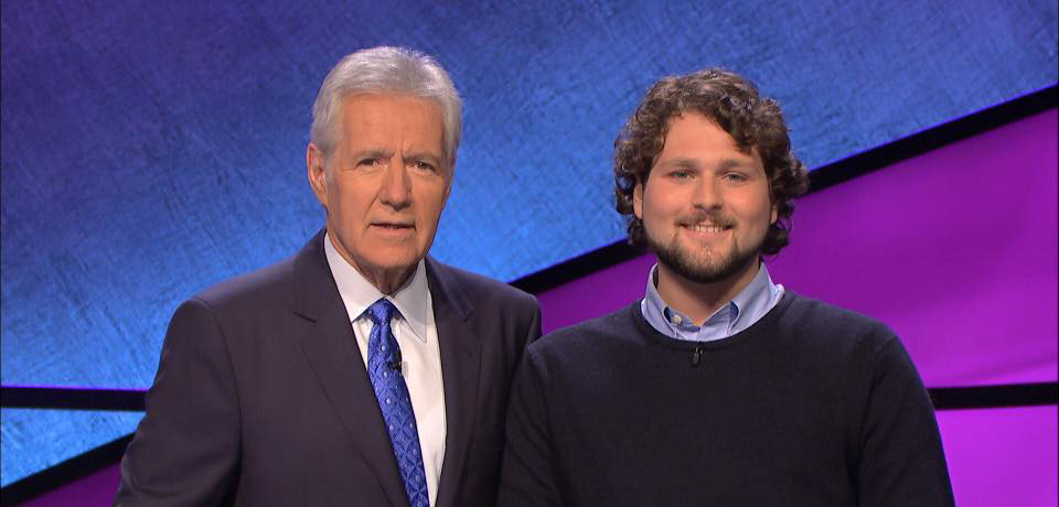 Alex Trebek in a dark suit standing next to a smiling contestant with curly dark hair and a beard, set against the Jeopardy! stage background.