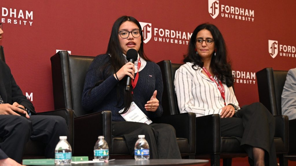 A teenage girl seated on a stage holding a microphone next to a woman in a white shirt at Fordham's Ignatian AI conference