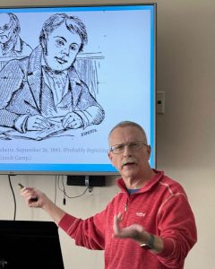 A man wearing glasses and a red pullover gestures while giving a presentation in front of a flat-screen television displaying a historical sketch of a person writing at a desk.