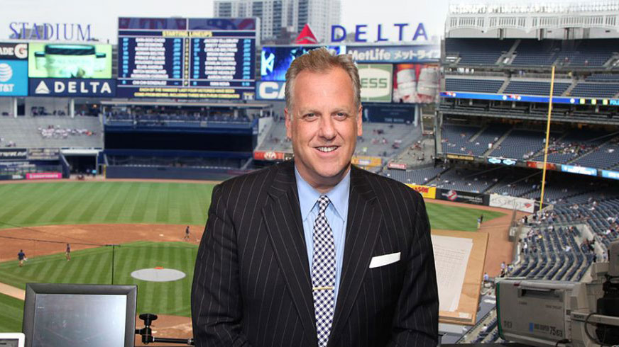 Broadcaster and Fordham graduate Michael Kay smiling in a suit in a broadcast booth at Yankee Stadium with a view of the field and center field scoreboard behind him.